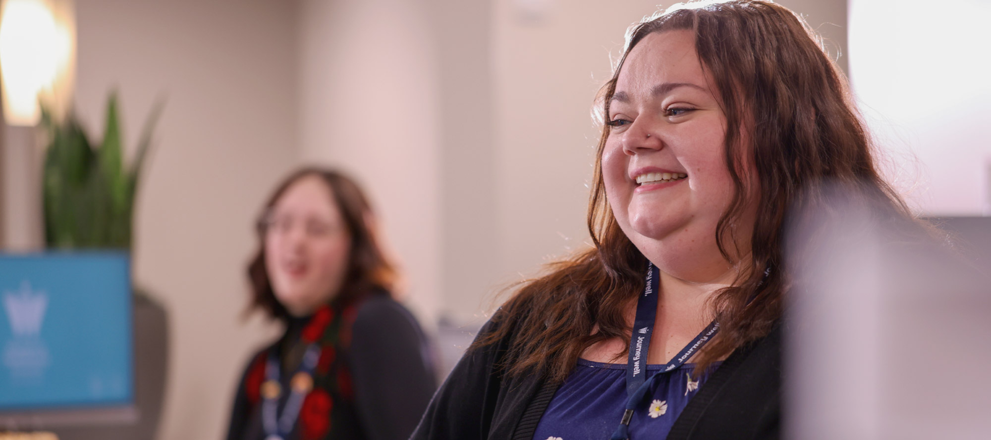 Clinic staff member wearing a badge smiles while standing in a healthcare office, with a colleague blurred in the background.