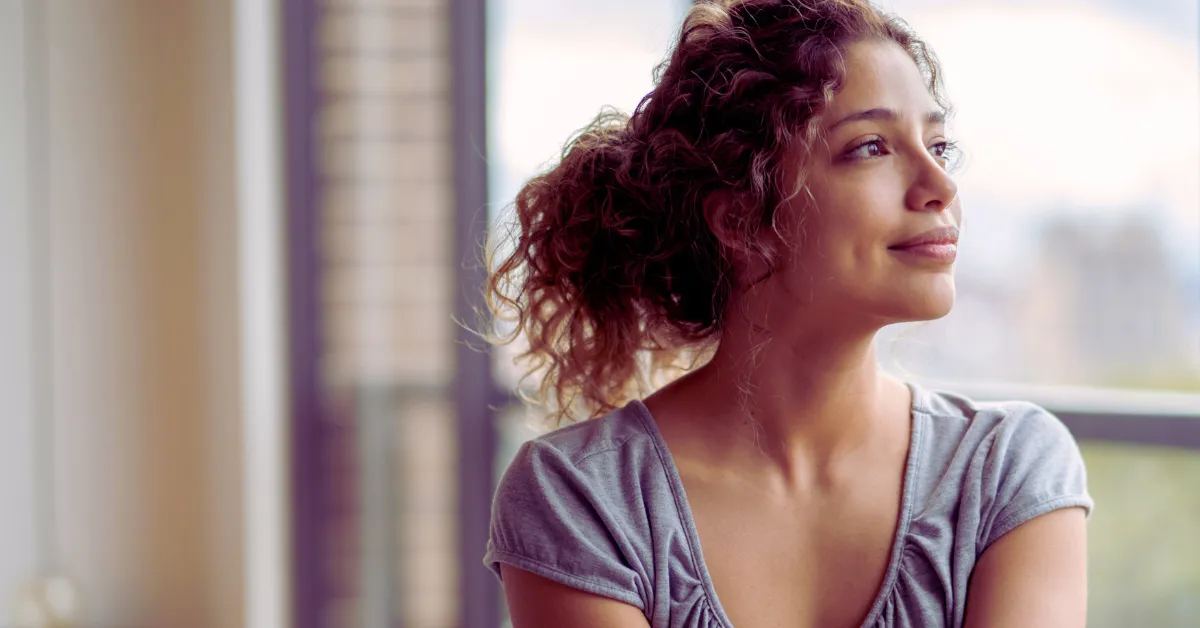 A woman looks out a sunlit window with a calm, reflective expression.