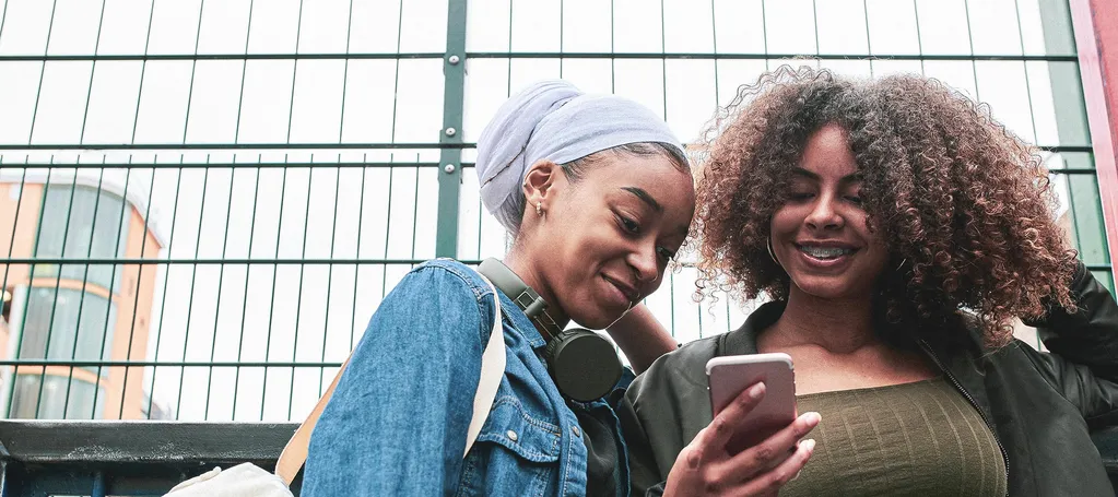 Two teenage girls standing against a metal fence, smiling as they look at a phone together.