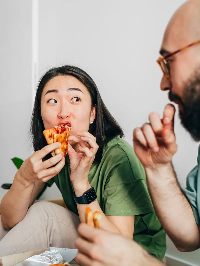 A woman eating pizza on the floor shares a playful moment with a man sitting beside her.