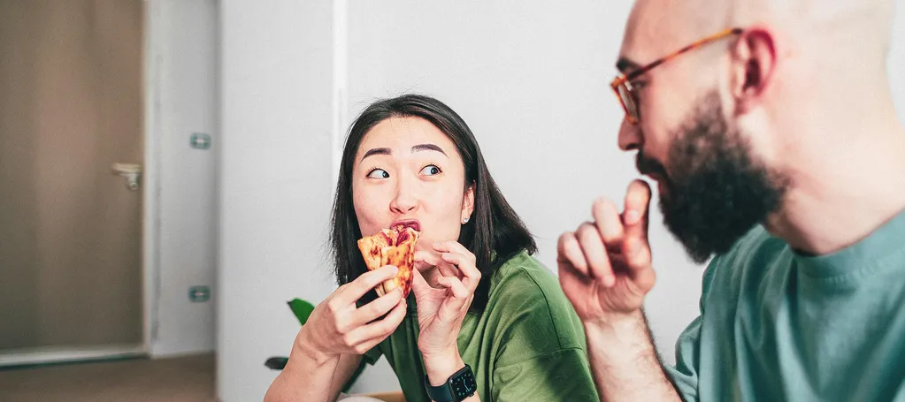 A woman eating pizza on the floor shares a playful moment with a man sitting beside her.
