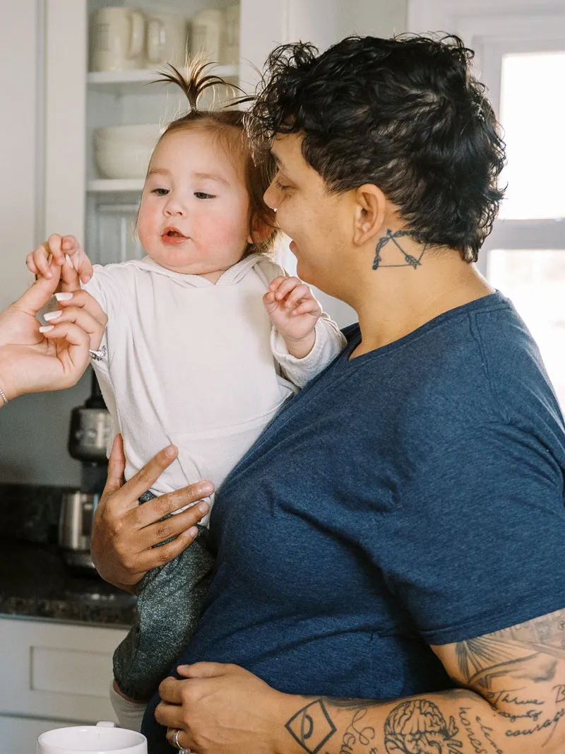 A parent holds a baby in a bright kitchen while another hands it something to eat.