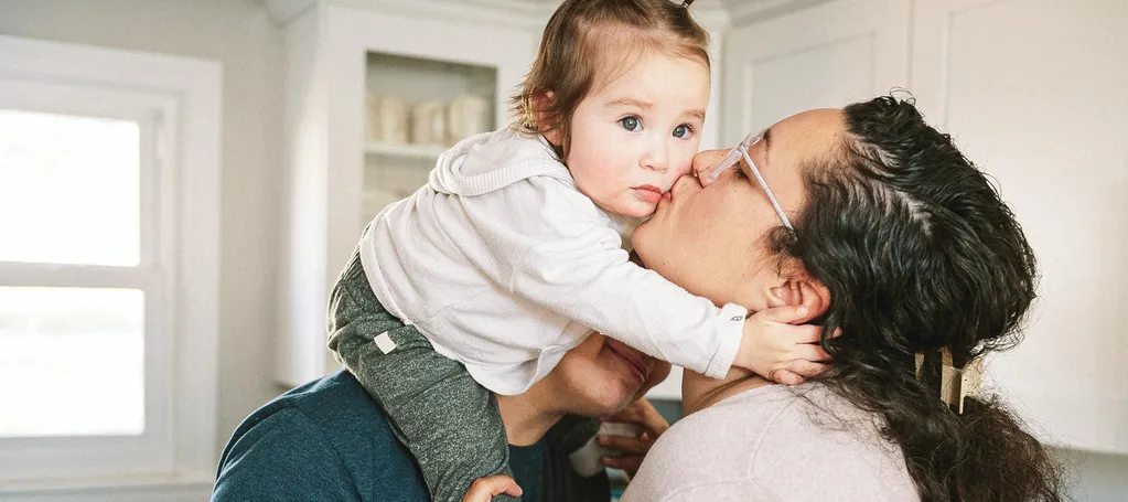 A parent lifts a baby for a kiss in a bright kitchen while another parent smiles behind them.