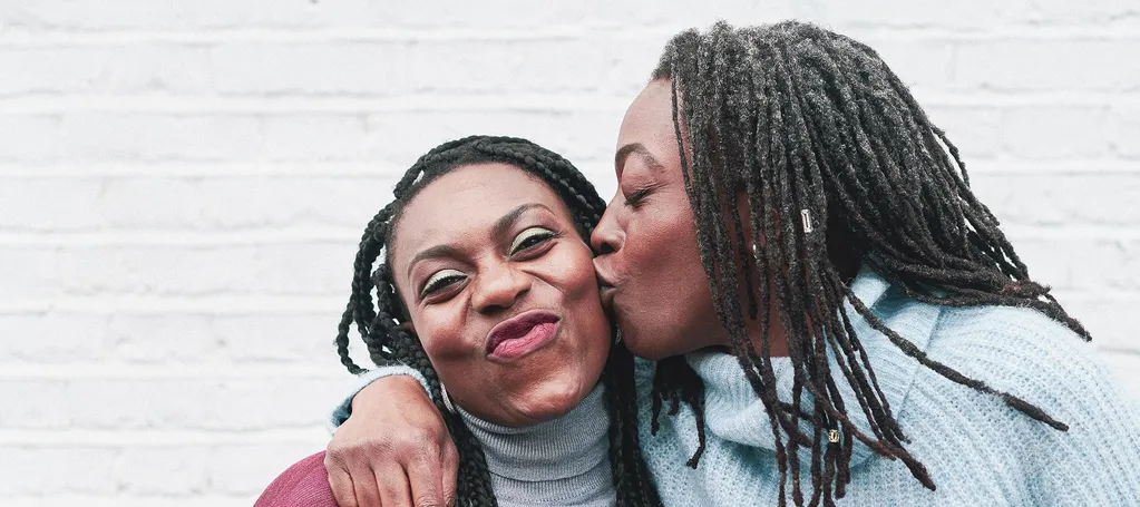 One woman hugs another and kisses her cheek in front of a white brick wall.