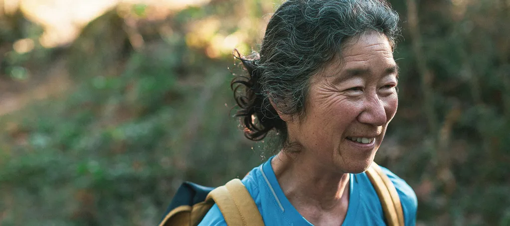 An older woman with a backpack smiles while walking outdoors on a forested trail.