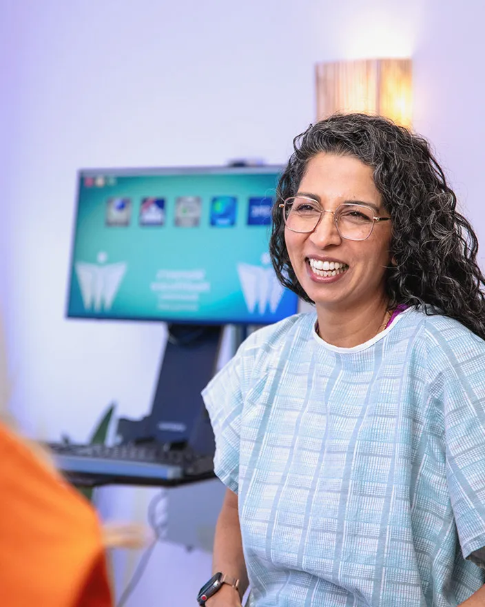Patient in a medical gown smiles while talking with a clinician in an exam room.