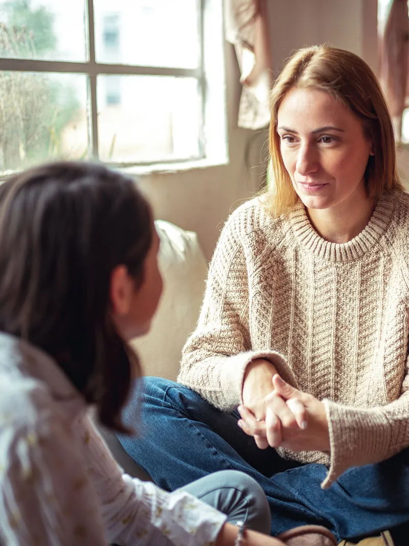 An adult woman sits cross-legged and listens attentively to a teenage girl in a softly lit room near a window.