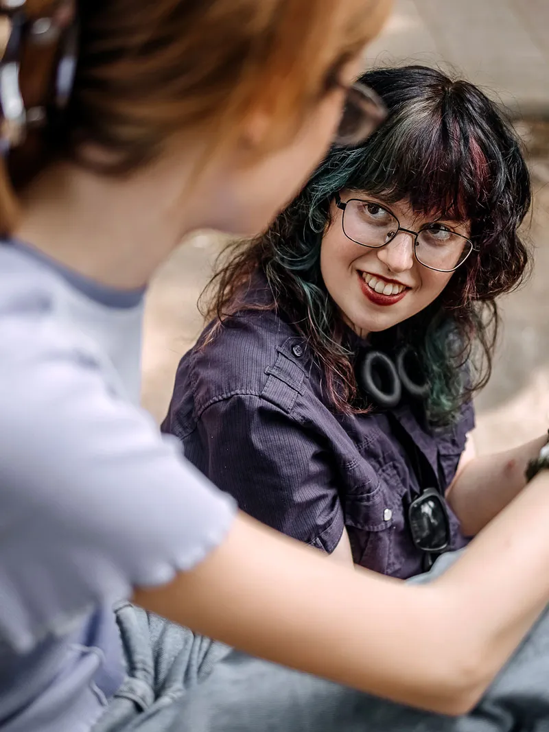 Two teenagers sit outdoors and talk, with one looking toward the other and smiling.