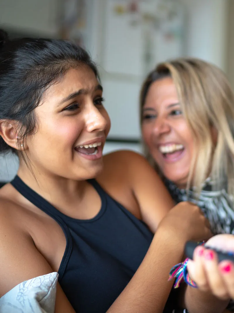 A teenage girl reacts playfully while sitting with an adult woman who is laughing beside her.