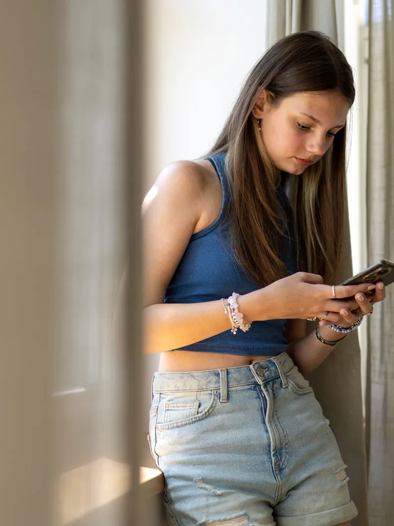 A teenage girl stands by a window and looks down at her smartphone.