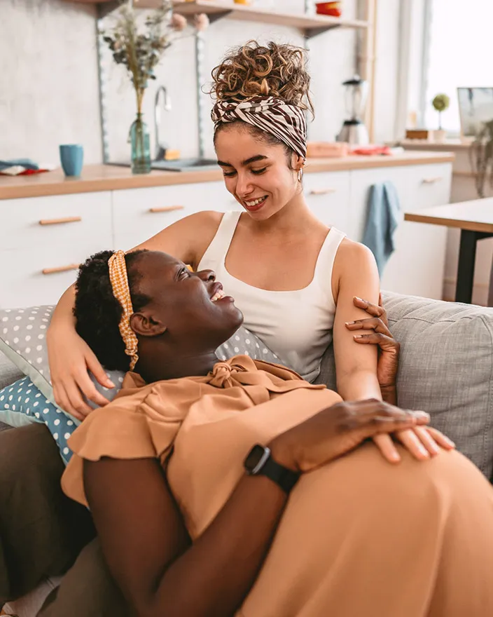 A pregnant woman reclines on a couch while her partner smiles down at her affectionately.