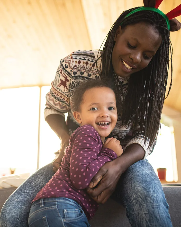 A woman hugs a young child on her lap indoors; both smile warmly.