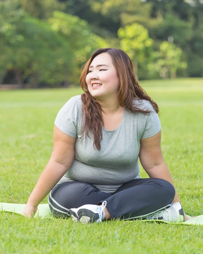 A woman sits cross-legged on a yoga mat in a park, smiling and looking off to the side.