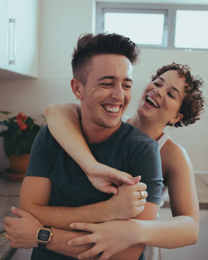 A couple laughs together as one wraps their arms around the other from behind in a kitchen.