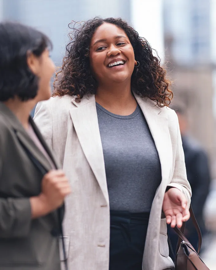 Two women walk and talk in a city setting, smiling during conversation.