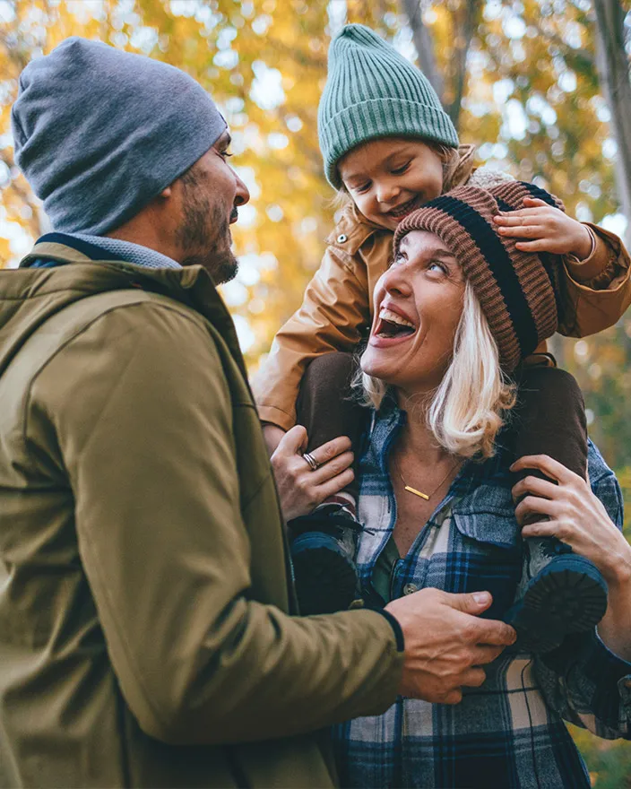 A parent stands with a child on their shoulders while another adult smiles nearby outdoors.