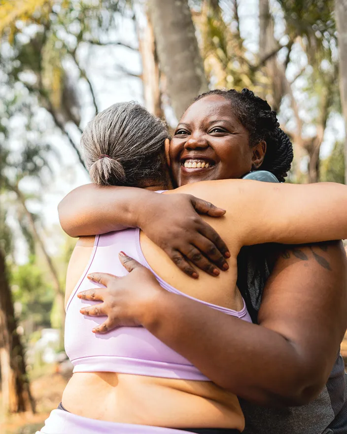 Two women share a warm hug outdoors, one smiling over the other’s shoulder.