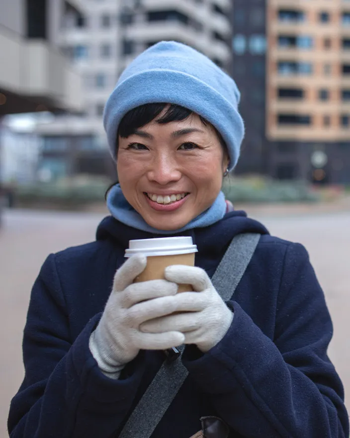 A woman in winter clothing holds a takeaway coffee and smiles at the camera.