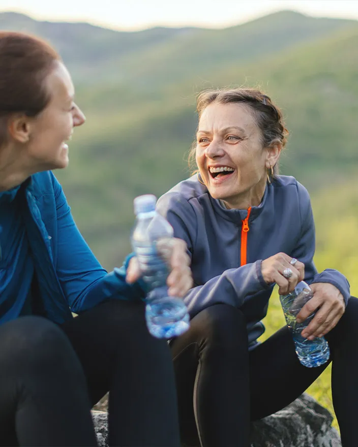 Two women sit on a hillside with water bottles, laughing during a hike.