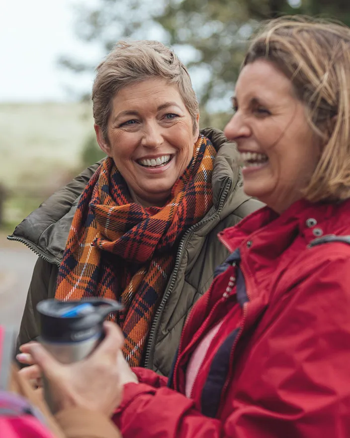 Two women outdoors in jackets laugh together while holding travel mugs.
