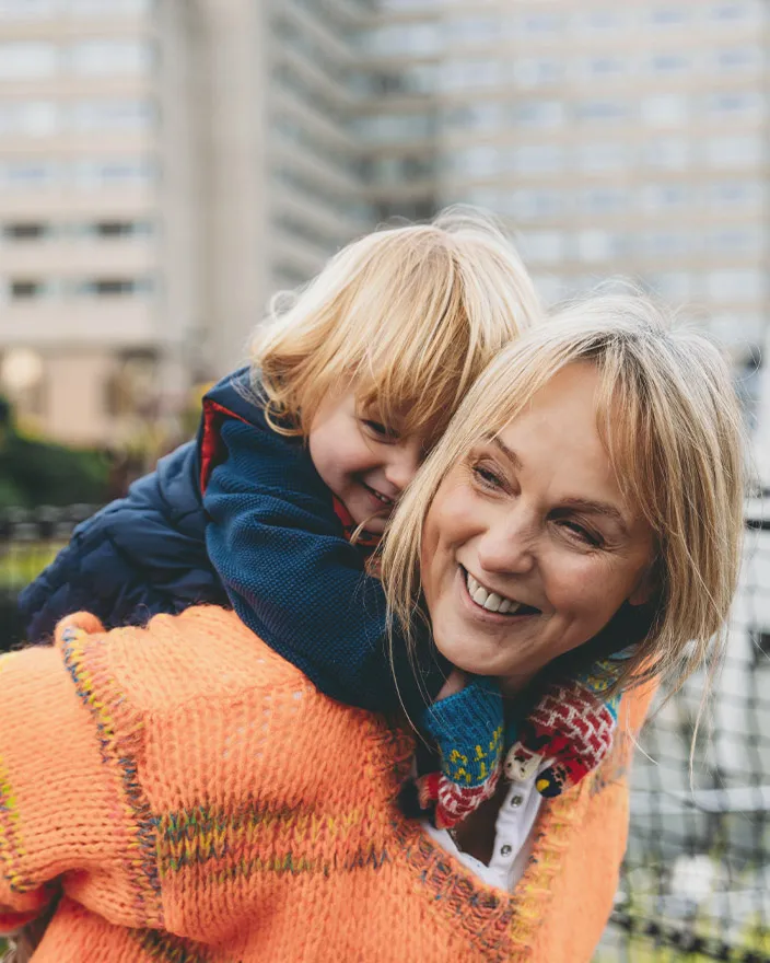 A woman carries a child on her back outdoors, both smiling playfully.