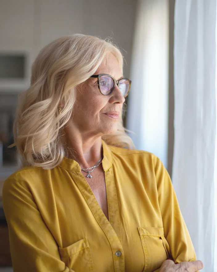 A woman stands by a window indoors, looking outside with a thoughtful expression.