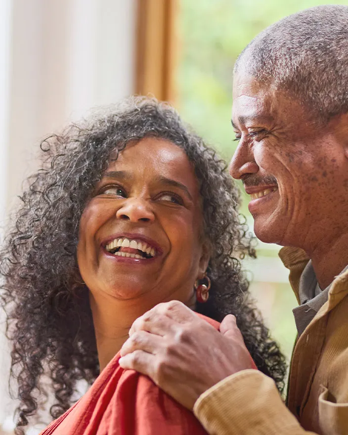 An older couple stand close together indoors, smiling and sharing a lighthearted moment.