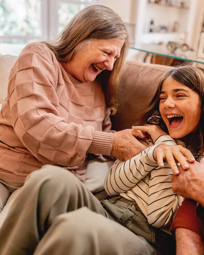 A grandmother and young girl laugh together on a couch while playfully interacting.