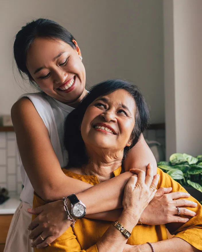A younger woman hugs an older woman from behind in a kitchen, both smiling warmly.