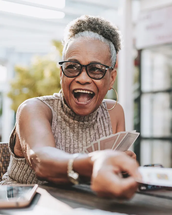 An older woman laughs while playing cards at a table, reaching forward with a playful expression.