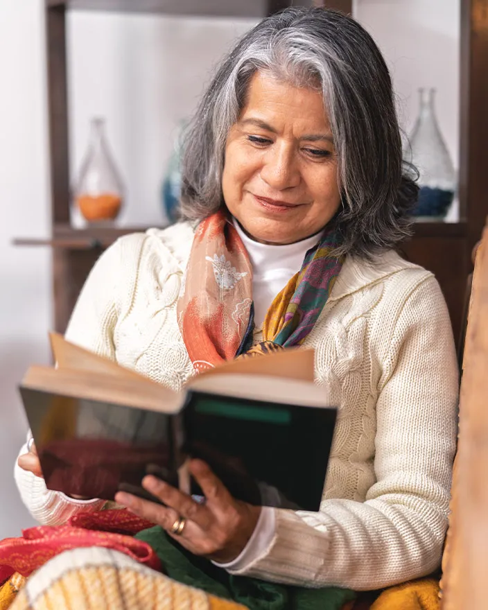 A woman sits comfortably indoors, smiling gently while reading a book.