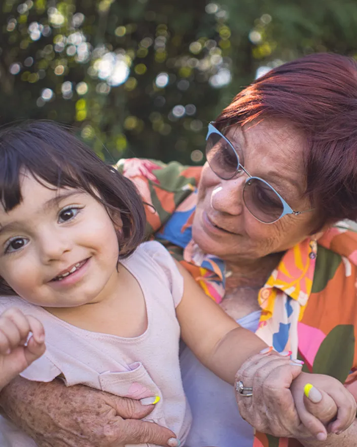 An older woman holds a young child outdoors, both smiling and engaged with each other.