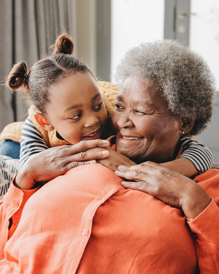 A grandmother and young child share a warm hug, smiling at each other indoors.