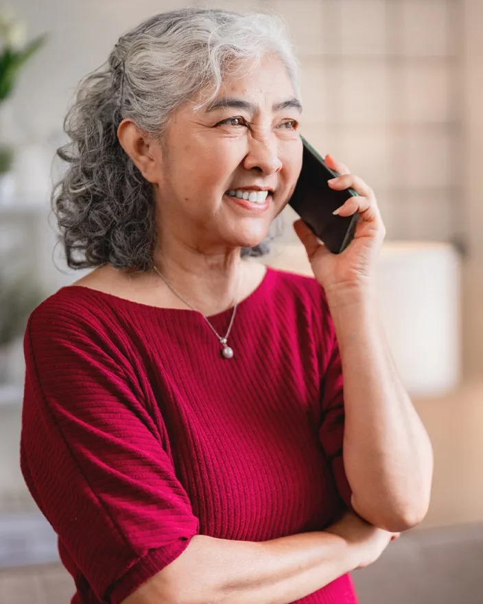 An older woman smiles while talking on a phone indoors.
