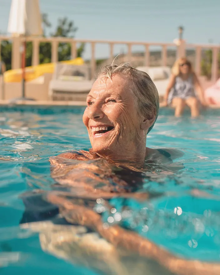 An older woman smiles while swimming in a pool on a sunny day.