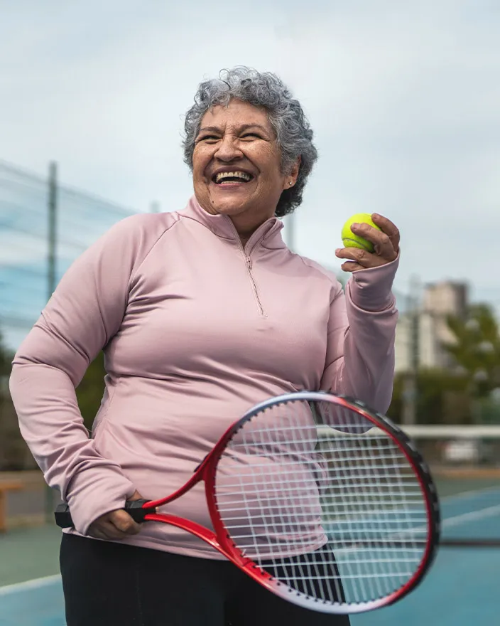 A woman stands on a tennis court holding a racket and ball, smiling confidently.
