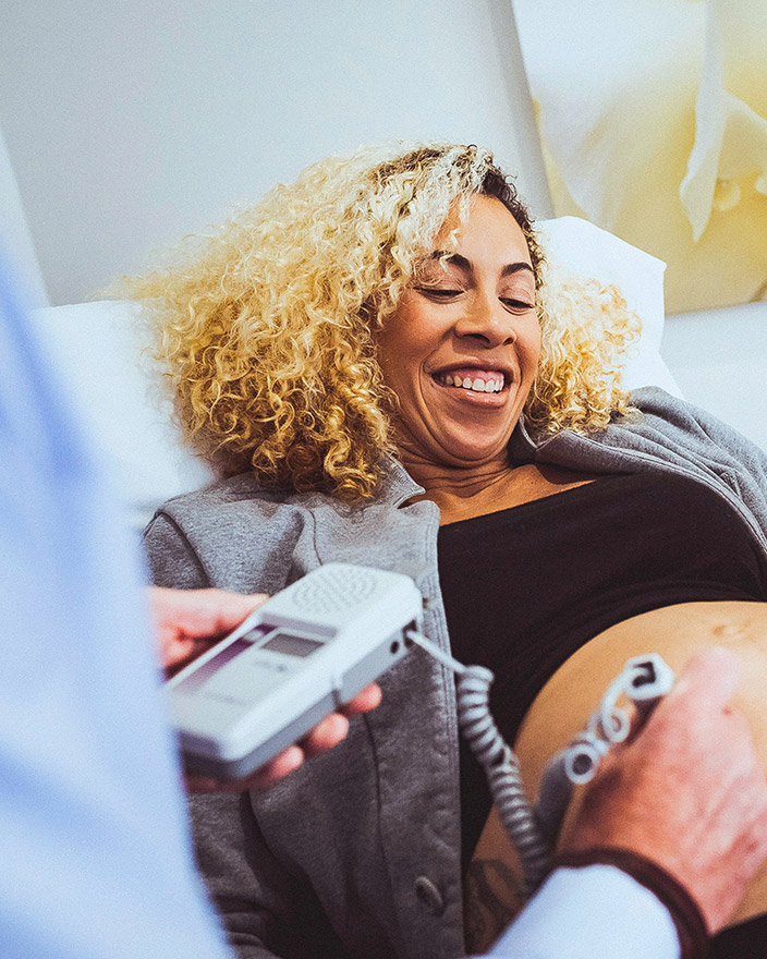 A pregnant patient smiles during a prenatal visit as a clinician checks the baby’s heartbeat with a monitor.